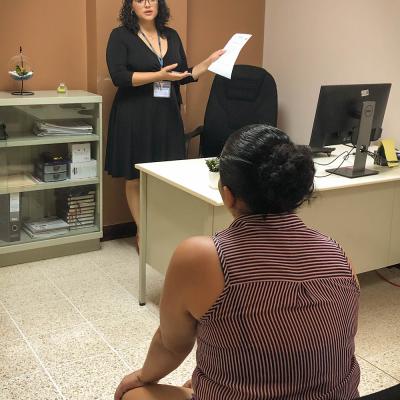 Señora de pie, conversa y le muestra un documento  a una mujer, ellas se encuentran en una oficina